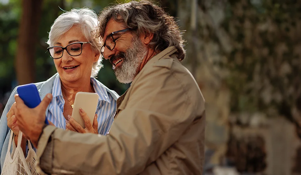 homem e mulher cerca de 60 anos mostrando celular um para outro em ambiente externo