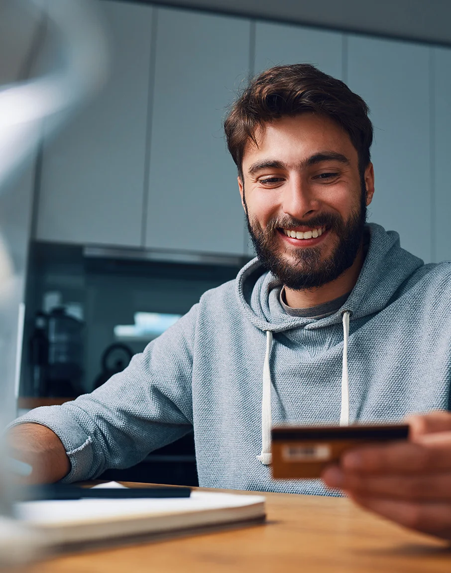 Homem jovem sorrindo usando moletom azul em mesa de casa com cartão de crédito em mãos