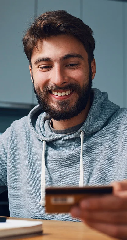 Homem jovem sorrindo usando moletom azul em mesa de casa com cartão de crédito em mãos